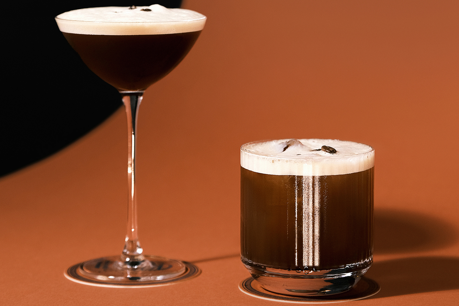 Studio shot of two coffee-based cocktails in coupe and rocks glasses with creamy foam on warm brown surface, Minimal dark background emphasizing rich espresso tones, Modern mixology and beverage design concept for bar menu and branding.